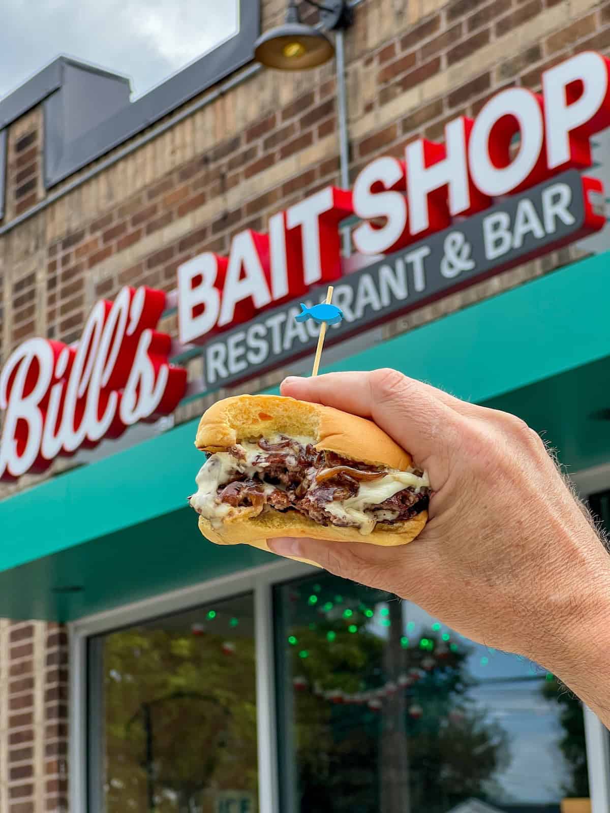 Hand holding a sandwich in front of Bill's Bait Shop storefront.
